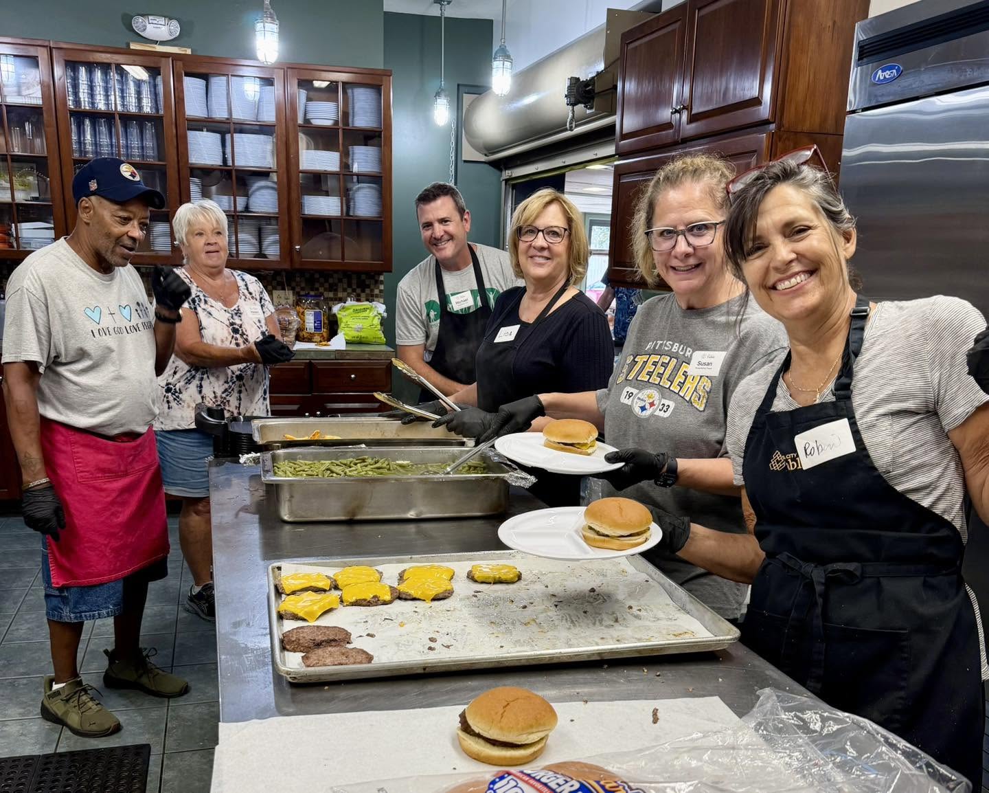 Steel City Table volunteers smiling behind the serving line, plating cheeseburgers and green beans